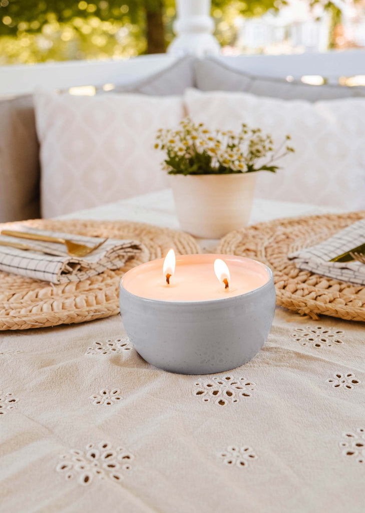 A lit The Bloom Candle from Mimi & August, featuring two wicks, sits on a lace tablecloth adorned with a dining setting that includes plates, napkins, and a small potted plant in the background.