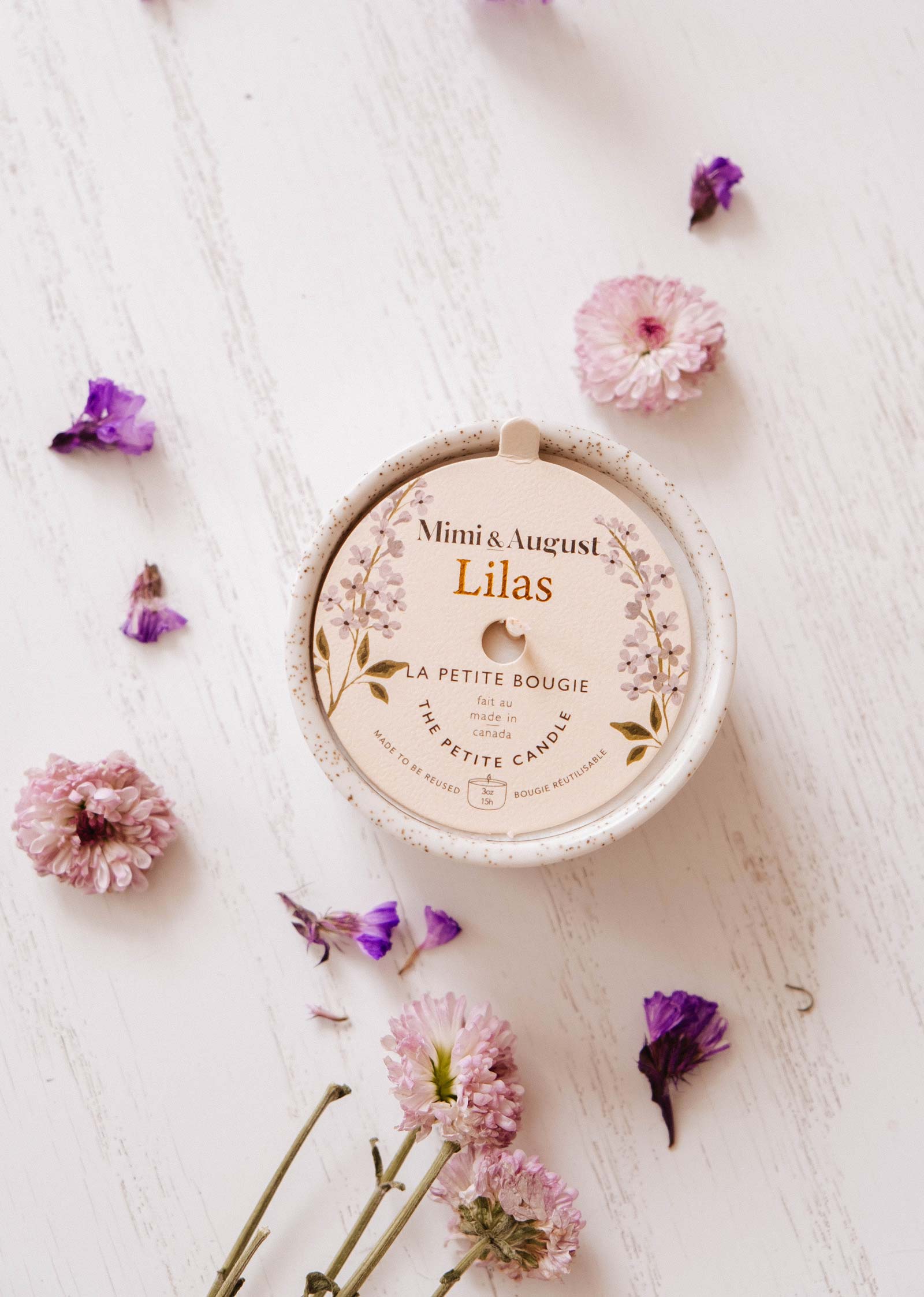 A Mimi & August Lilac - Reusable Candle sits on a white wooden surface, ideal for aromatherapy, surrounded by scattered pink and purple flowers.