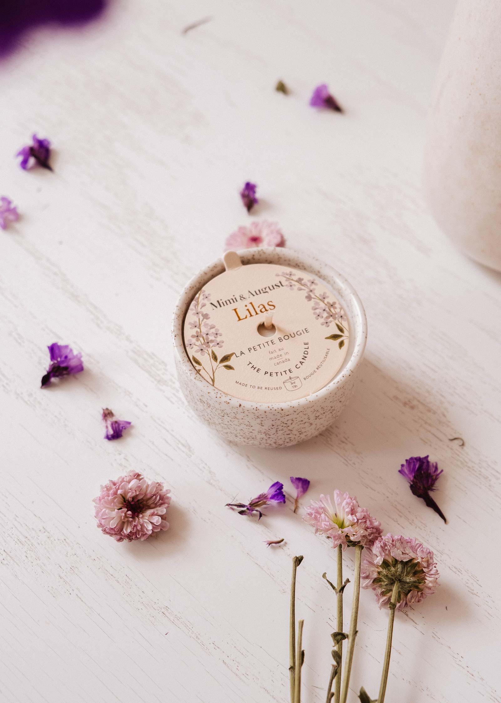 The Lilac - Reusable Candle by Mimi & August rests on a white wooden surface, surrounded by pink and purple flowers, bringing elegant home decor and soothing lilac fragrance to the space.