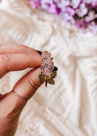 A hand holds the Mimi & August Lilac Enamel Pin, featuring gold tone brass with green leaves; a soft fabric and purple flowers are blurred in the background.
