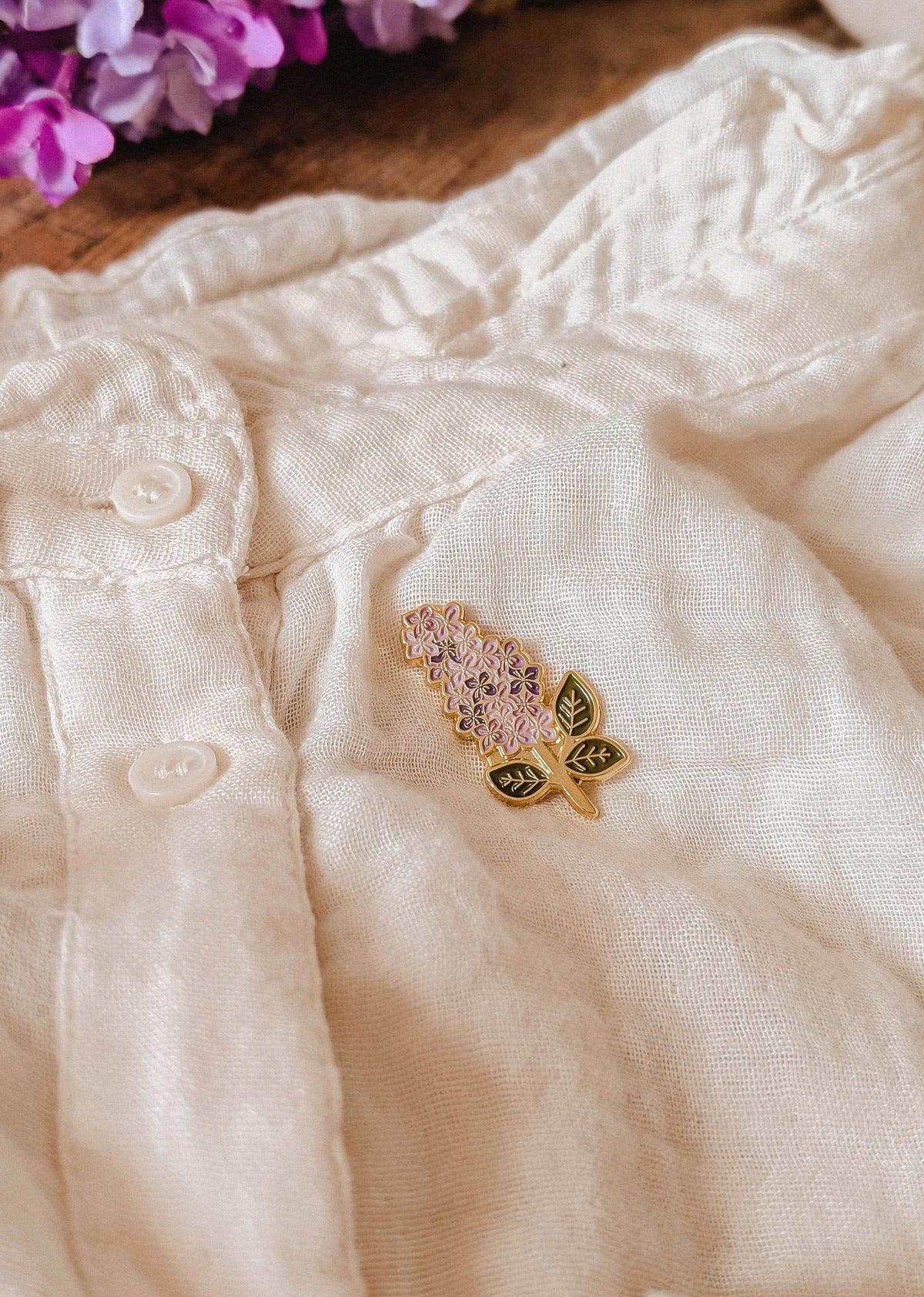 The Mimi & August Lilac Enamel Pin in gold tone brass is attached to a cream linen garment with buttons, with lilac flowers visible in the upper left corner.