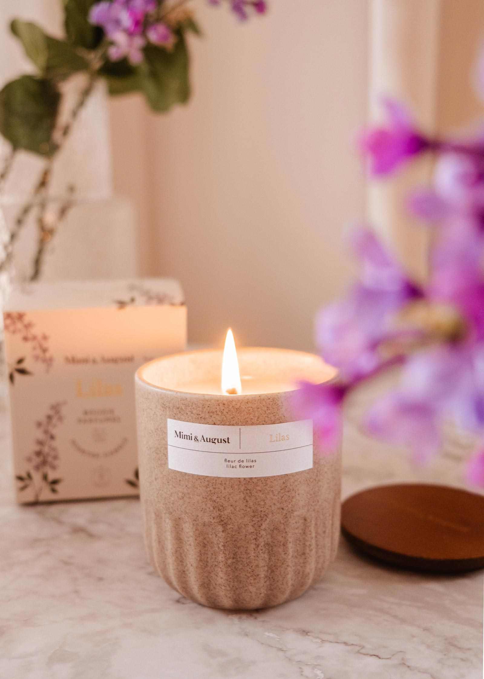 The Lilac - Reusable Candle by Mimi & August in a beige ceramic jar sits on marble, bringing aromatherapy and elegance to your home, with its box and purple flowers as a stylish backdrop.