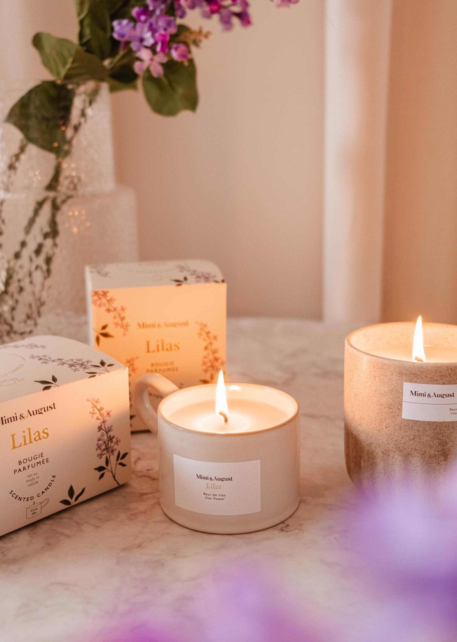 Two lit Mimi & August Lilac - Reusable Candles labeled "Lilas" sit on marble, with a vase of purple flowers in the background—ideal for adding soothing aroma and elegant home decor to your space.