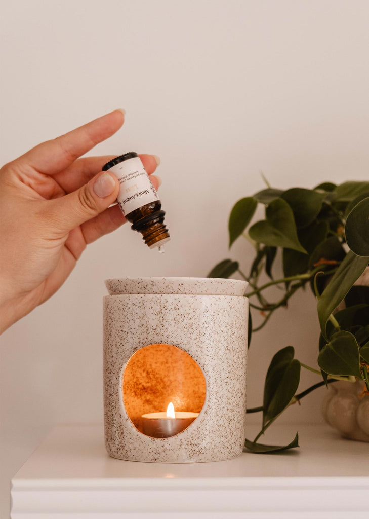 A hand is holding a small bottle of Scented Oil - Lilac by Mimi & August, adding a few drops into an aroma diffuser with a lit tealight candle inside. A green plant is visible in the background, enhancing the serene atmosphere with lilacs' soothing scent.