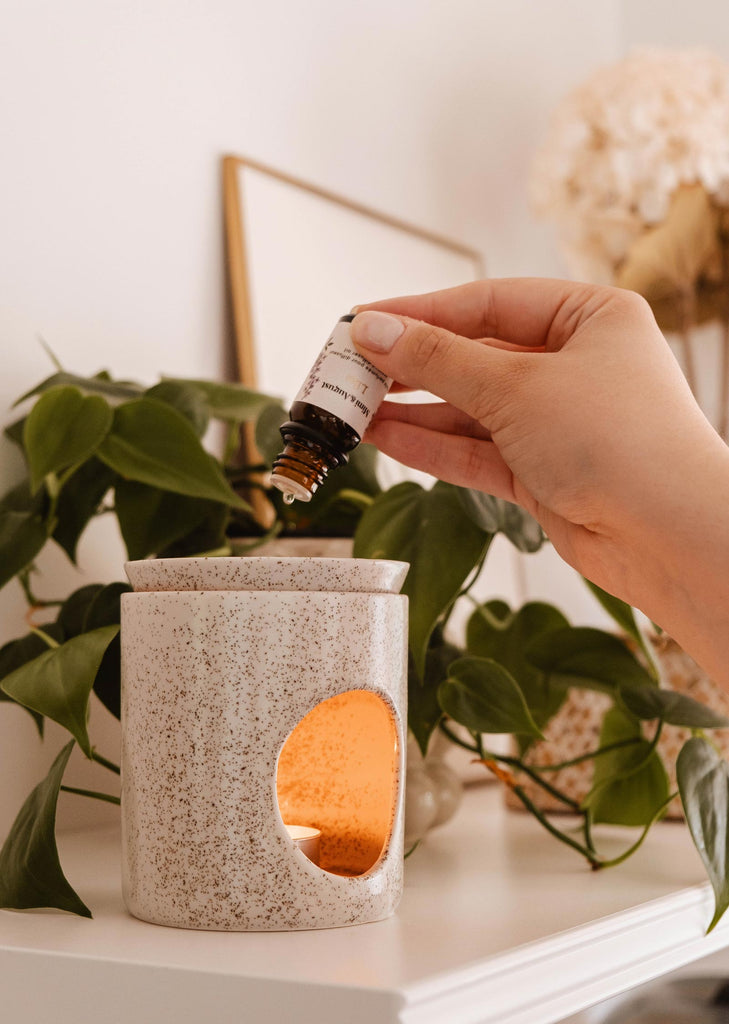 A hand is shown adding a few drops of Mimi & August Scented Oil - Lilac into a lit ceramic diffuser, set against a backdrop of green leaves and blurred decor items, creating an ambiance reminiscent of lilacs in spring.