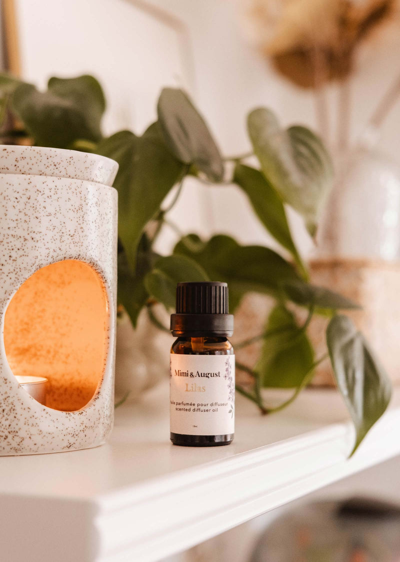 A small bottle labeled "Mimi & August Scented Oil - Lilac" is placed on a shelf beside a white ceramic oil diffuser with a lit candle inside, releasing its calming and soothing lilac scent. In the background, green leaves and neutral decor are visible.