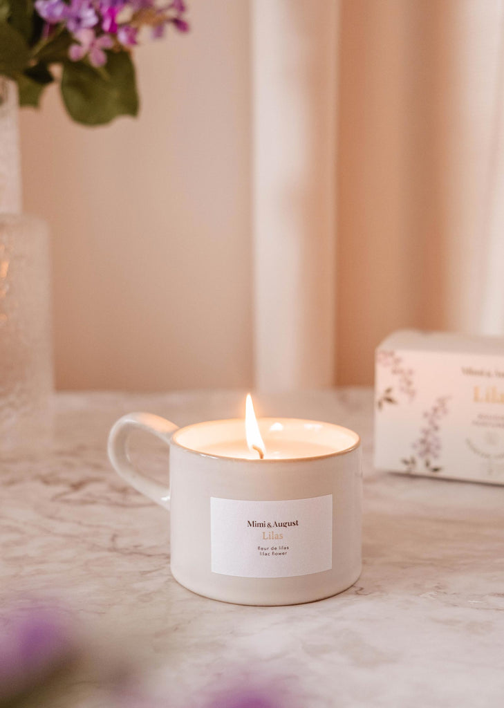 A Lilas scented candle in a white container with the label "Mimi & August" on a marble surface, surrounded by soft pink and purple hues.