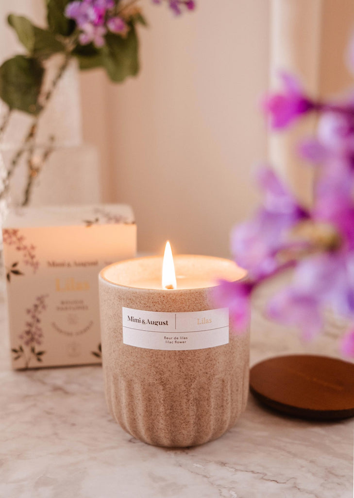 Une bougie parfumée Lilas de Mimi & August allumée dans un support décoratif sur une surface en marbre, avec une composition florale et un emballage en arrière-plan.