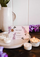 A Mimi & August Lilac Tealight Candle Set rests on a ceramic plate with four soy tealights—one lit—surrounded by lilac flowers and a white pitcher in the background.