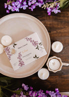 The Mimi & August Lilac Tealight Candle Set features a floral lilac box on a plate, surrounded by natural soy wax tealights, a ceramic holder, and purple flowers on a wooden surface.