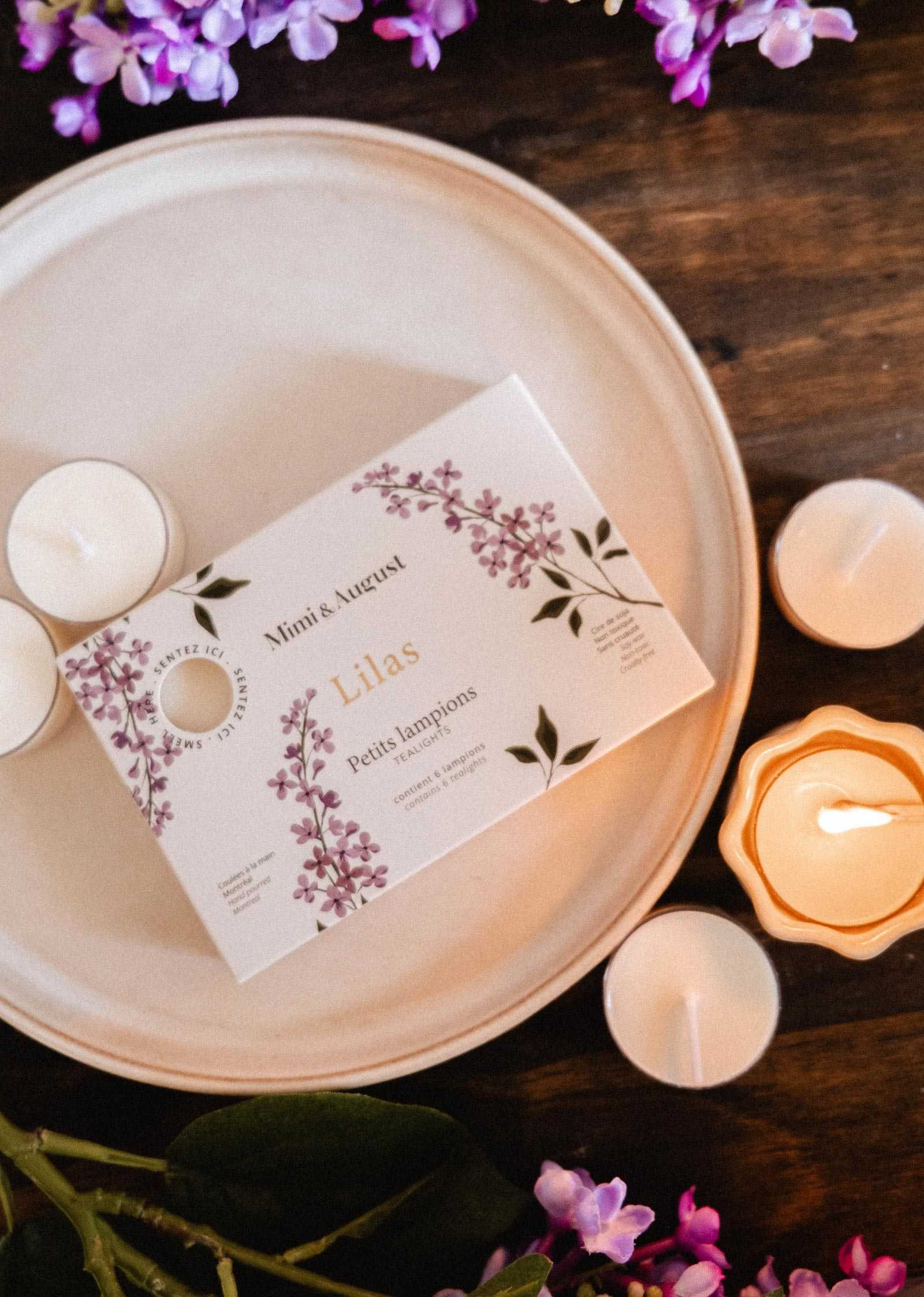 The Lilac Tealight Candle Set by Mimi & August, in a floral-designed box, is displayed on a plate with soy tealights and purple flowers arranged on a wooden surface.