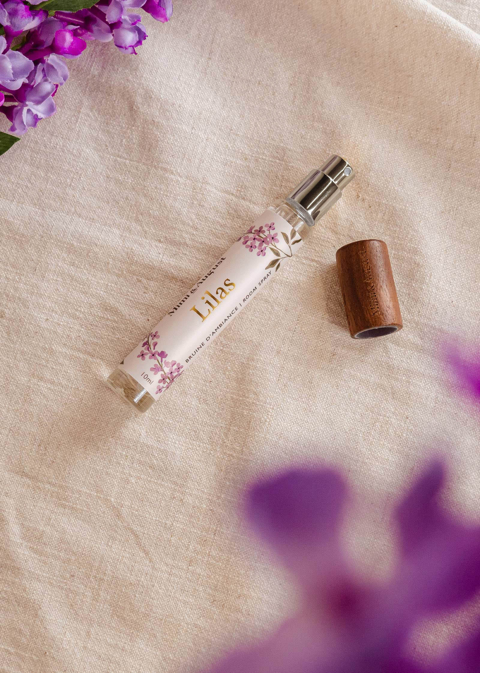 Un flacon pulvérisateur de Mimi & August Room spray - Lilas est posé sur une surface en tissu, avec un bouchon en bois à côté. Des fleurs violettes se trouvent en haut à gauche et en bas à droite de l'image, dégageant un parfum de lilas.