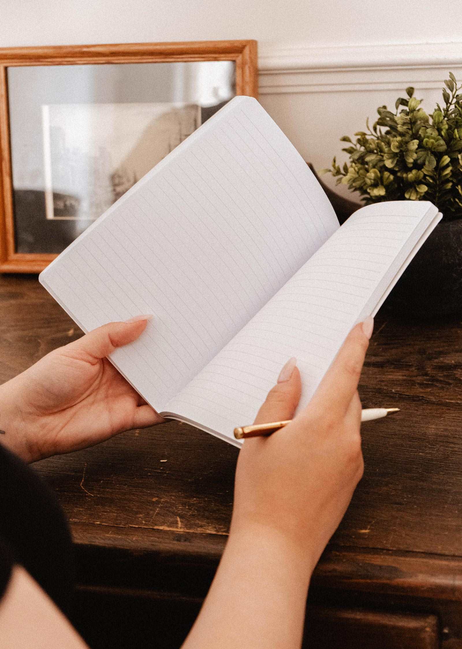 A person holds the open Fields of Flower floral notebook by Mimi & August in one hand and a pen in the other.