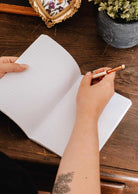 A person holds a pencil above an open Fields of Flower floral notebook by Mimi & August, beside a potted plant and a small frame.