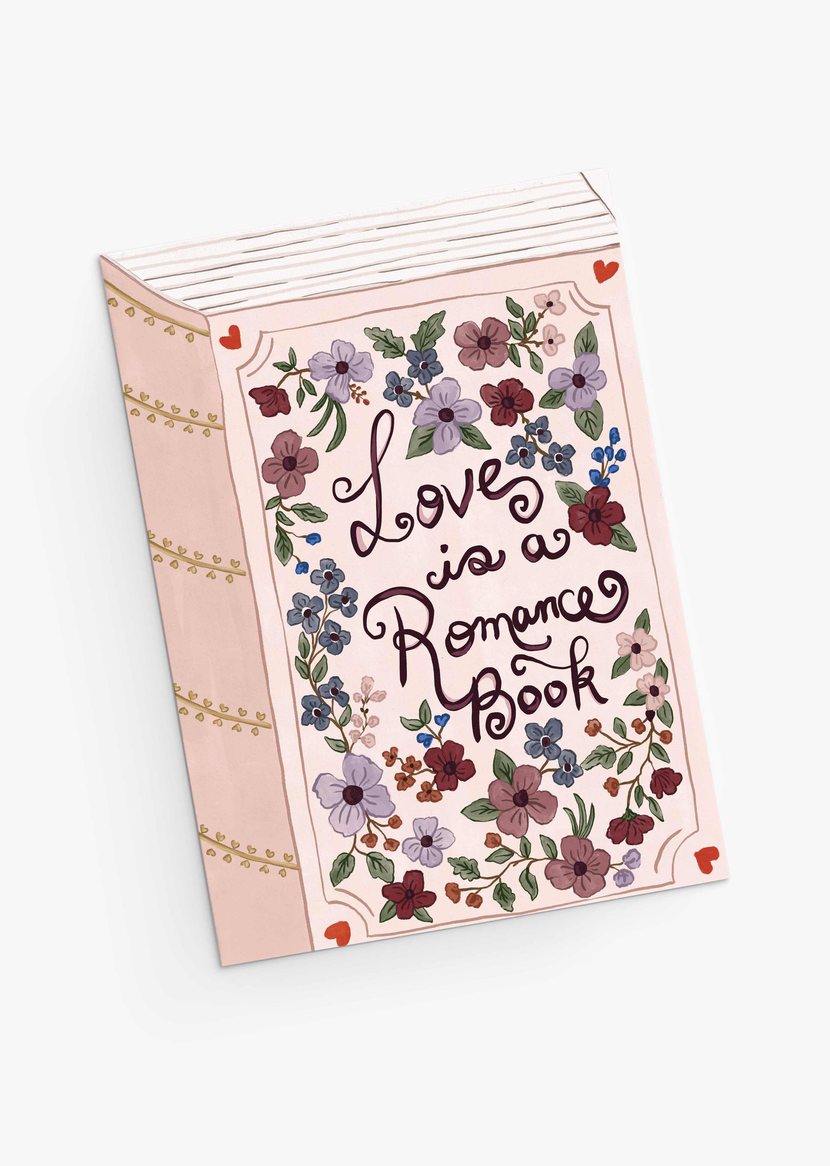 The Mimi & August "Love is a Romance Book" Greeting Card features a pink cover with floral illustrations and cursive text.