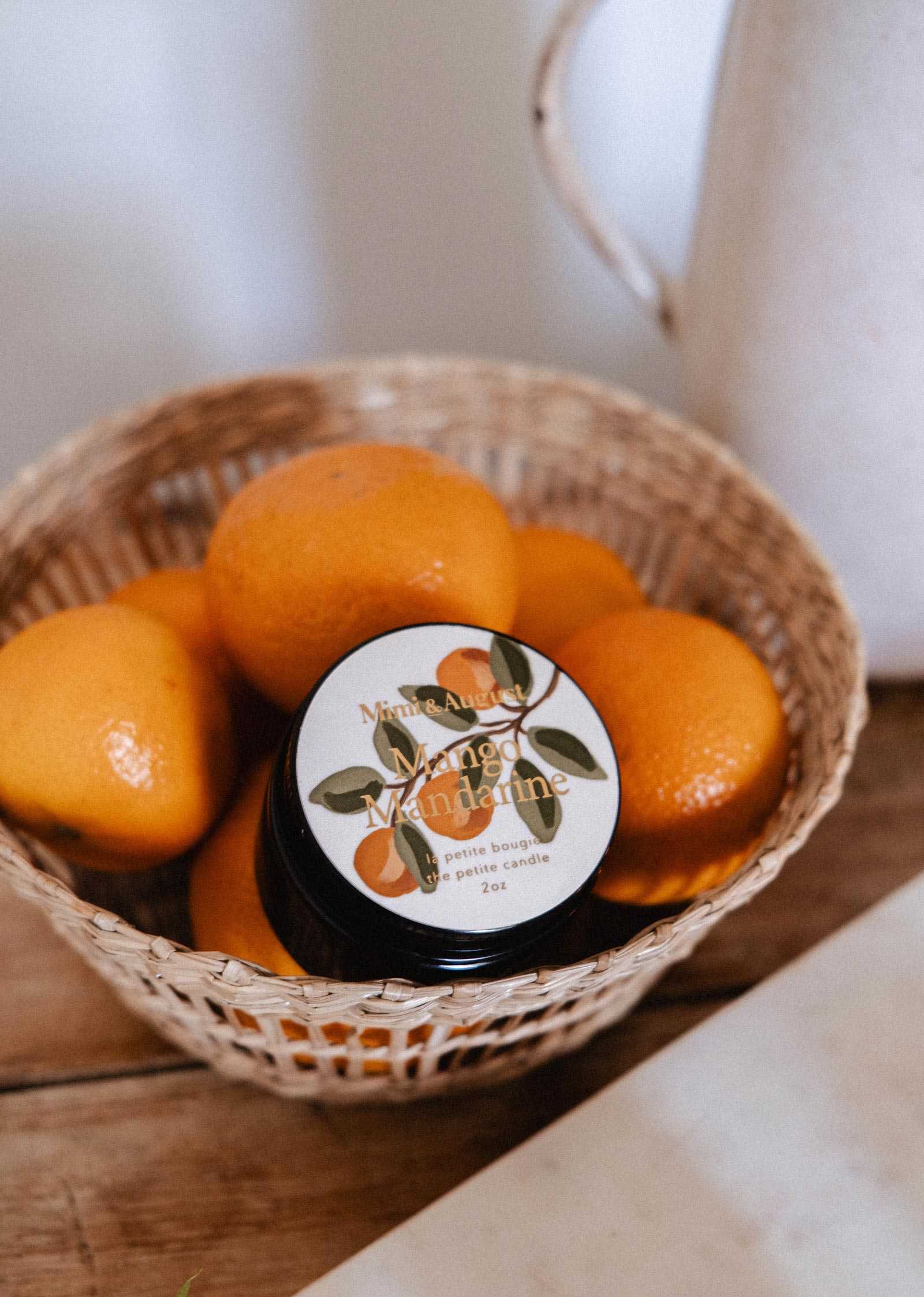 A small woven basket filled with mandarins and a Mimi & August Mango Mandarine – 2 oz Soy Candle sits on a wooden surface.