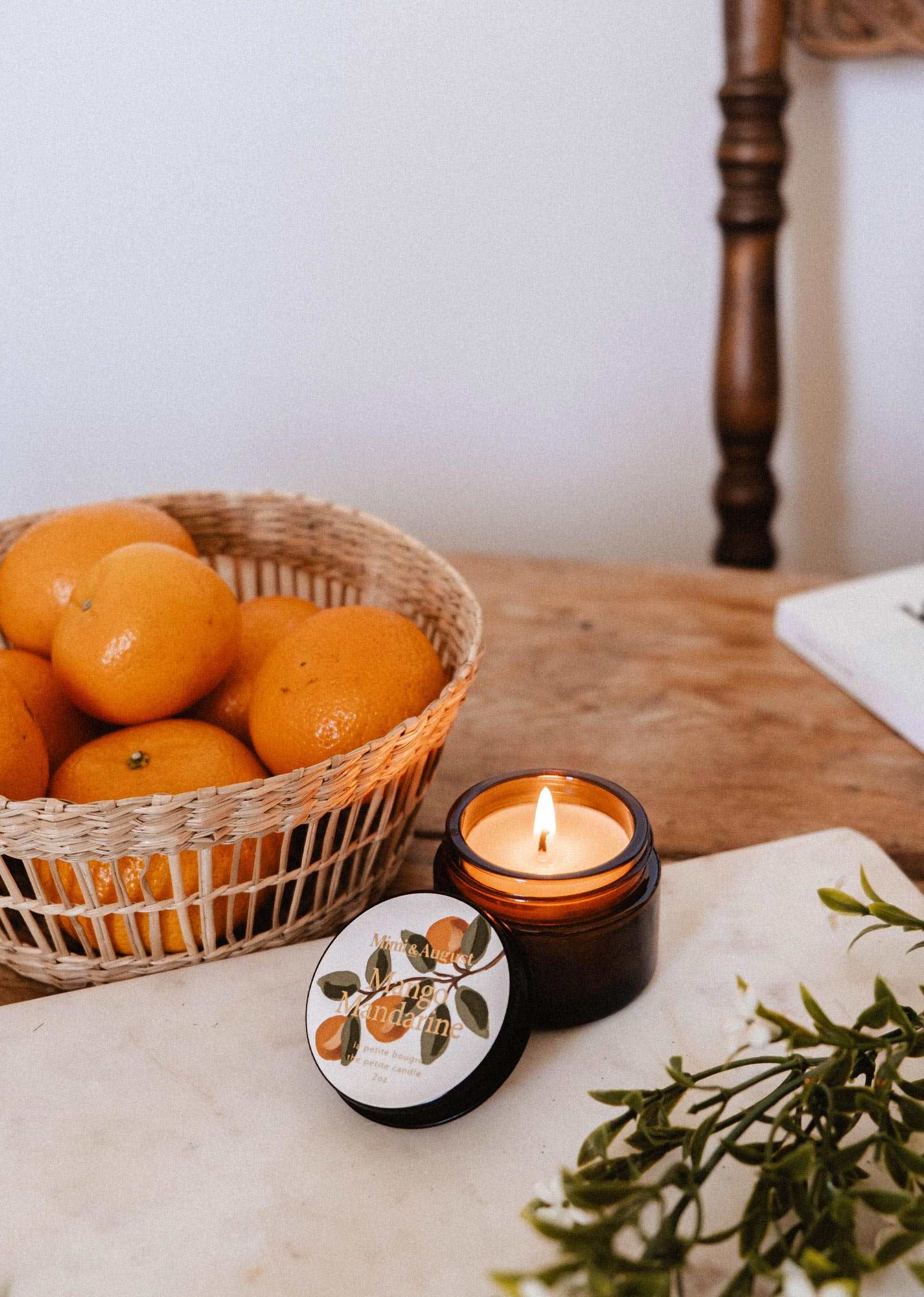 The Mimi & August Mango Mandarine – 2 oz Soy Candle in a brown jar rests beside its lid, a basket of oranges, and greenery on a wooden table.