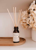 The Gift March - Free Reed Diffuser by Mimi & August, a brown glass bottle labeled "Eucalipto" with reed sticks on a wooden tray, provides continuous fragrance next to white hydrangeas on a shelf.