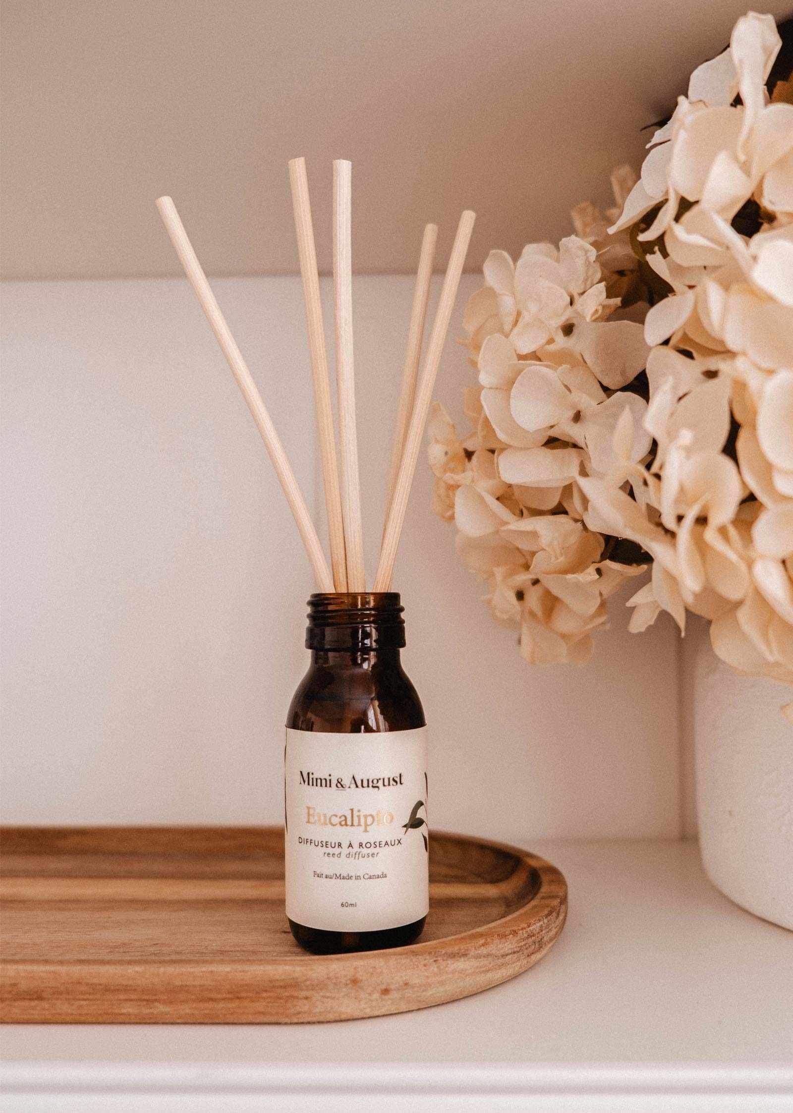 The Gift March - Free Reed Diffuser by Mimi & August, a brown glass bottle labeled "Eucalipto" with reed sticks on a wooden tray, provides continuous fragrance next to white hydrangeas on a shelf.