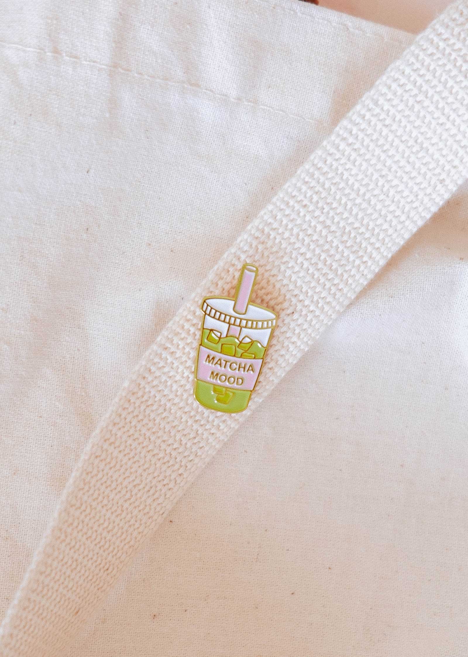 The Matcha Mood Enamel Pin by Mimi & August is a cute cup of matcha bubble tea with "MATCHA MOOD" text, attached to a white cloth bag—an ideal gift for any matcha lover.