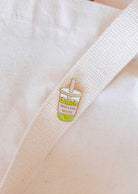 The Matcha Mood Enamel Pin by Mimi & August is a cute cup of matcha bubble tea with "MATCHA MOOD" text, attached to a white cloth bag—an ideal gift for any matcha lover.