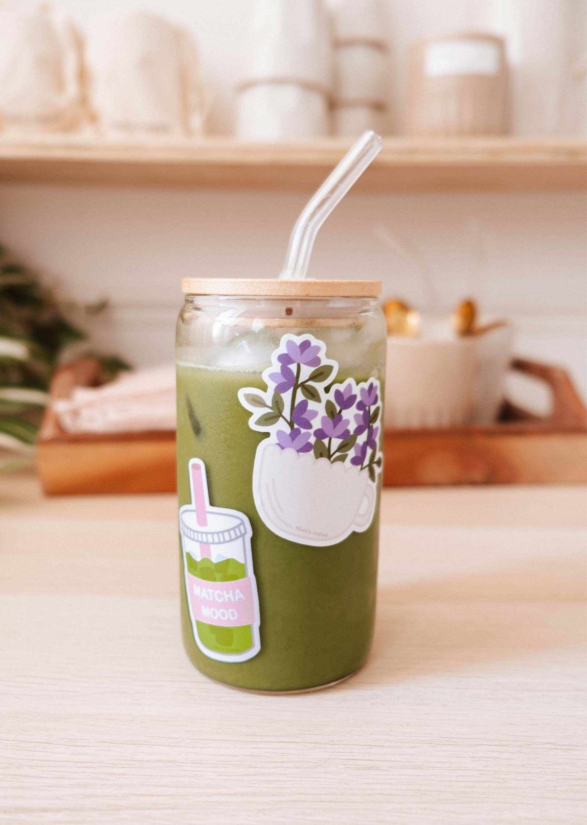 A glass jar of iced matcha latte with a bamboo lid, clear straw, and the Matcha Mood - Vinyl Sticker by Mimi & August sits on a light wooden table, with blurred kitchen items visible in the background.