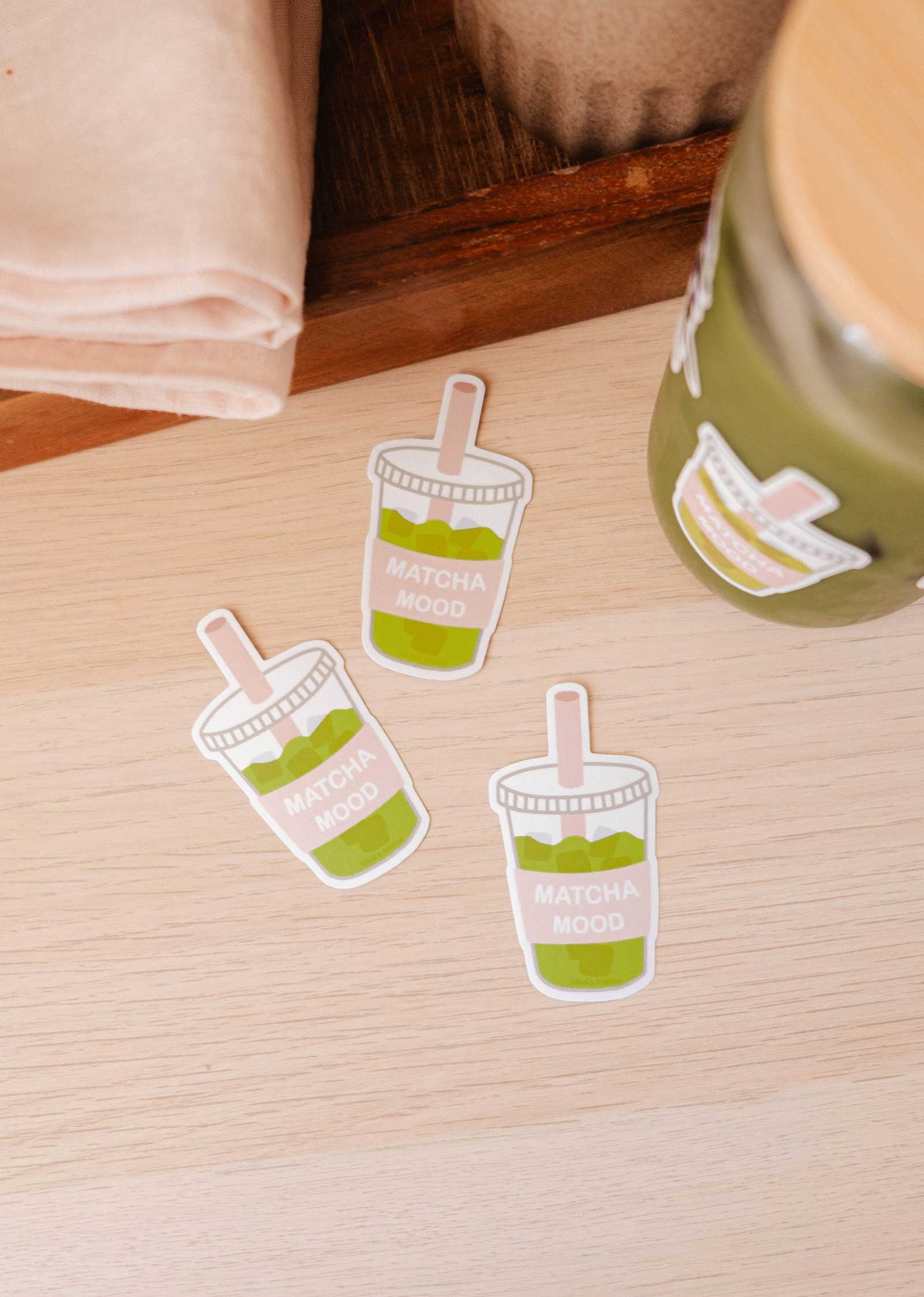 Three Mimi & August "Matcha Mood - Vinyl Sticker" durable iced matcha drink-shaped stickers rest on a light wooden surface beside a jar with the same sticker and a folded beige napkin.