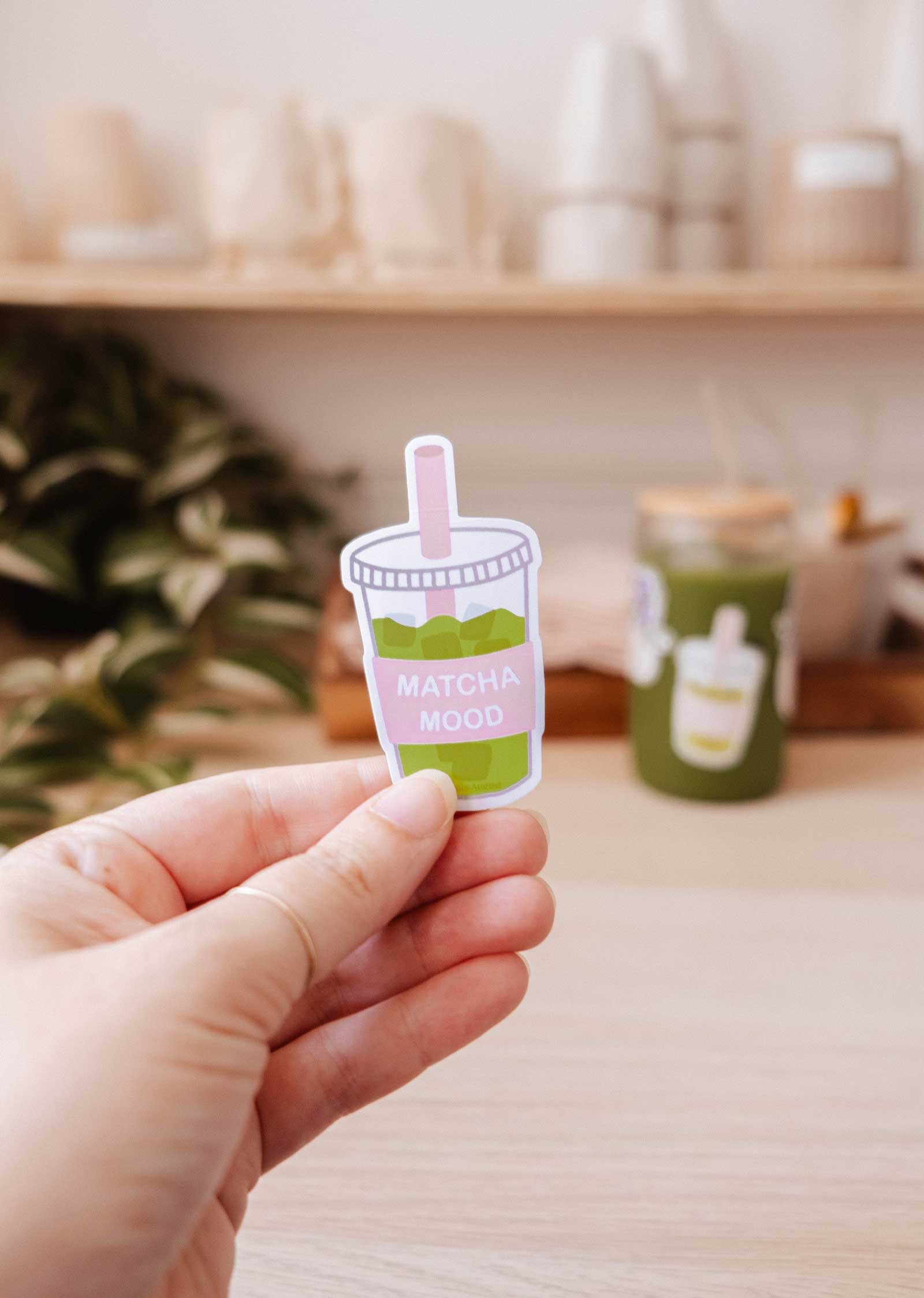 A hand holds the "Matcha Mood - Vinyl Sticker" by Mimi & August, featuring a cartoon iced matcha and the label "MATCHA MOOD," with a blurred green cup and shelves in the background.