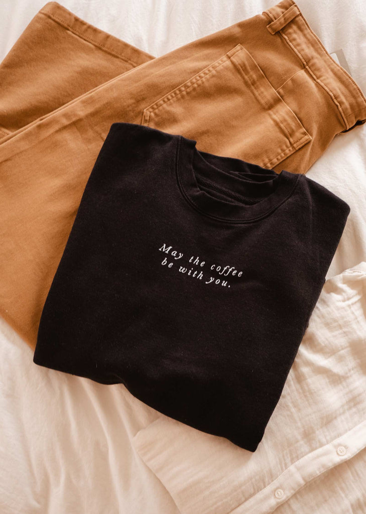 A comfortable May the coffee be with you Sweatshirt from Mimi & August laid on a bed with tan pants.