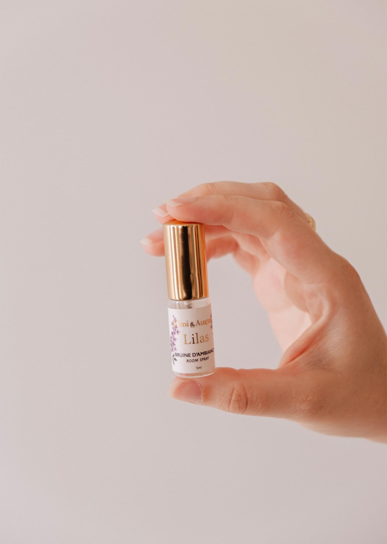 A hand holds a Mini Room spray - Lilas by Mimi & August, featuring a gold cap and label, against a plain light background—the perfect way to enjoy a delightful lilac fragrance.