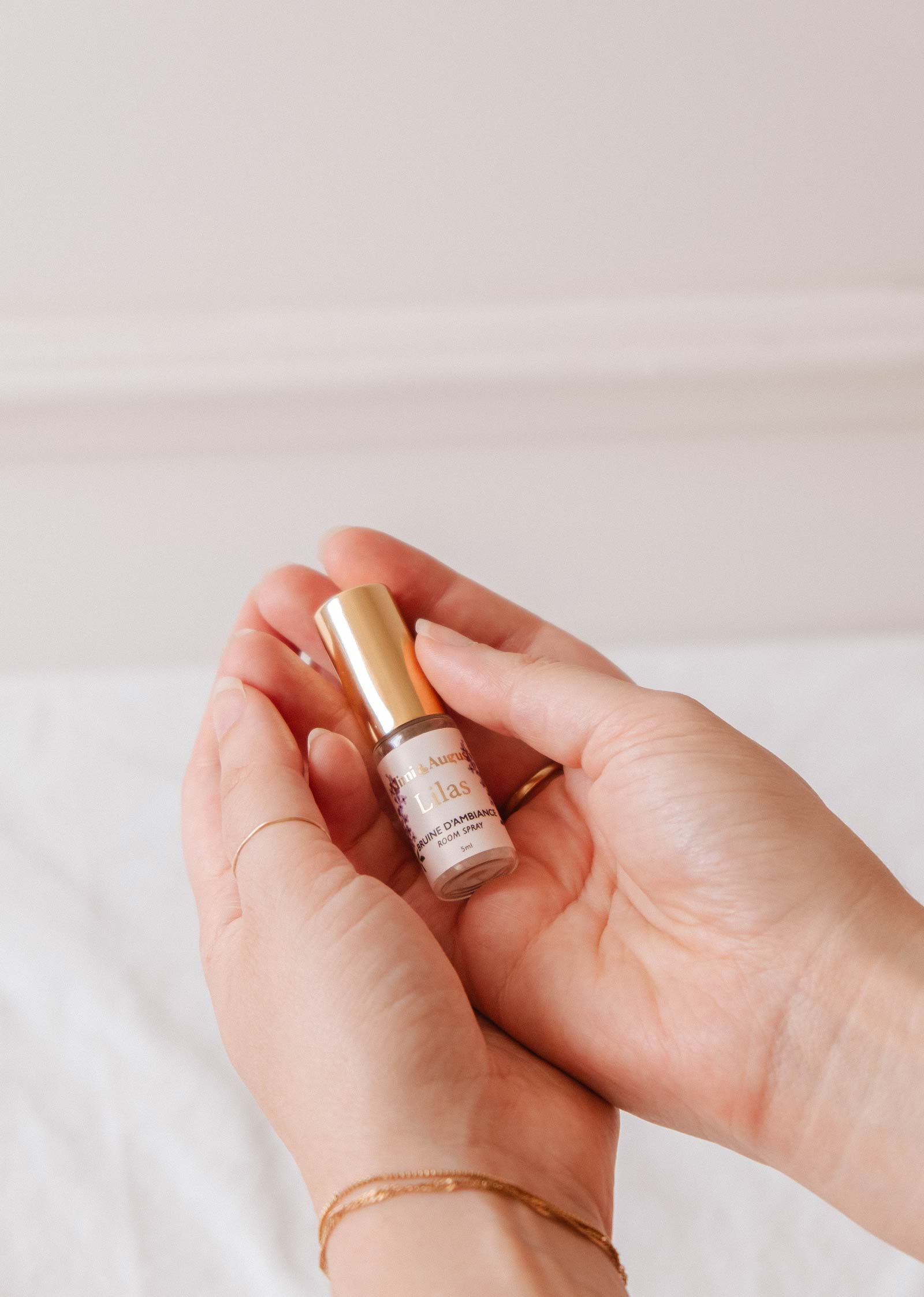 Deux mains tiennent un mini spray d'ambiance - Lilas de Mimi & August, dont le flacon à bouchon doré et le design élégant évoquent une ambiance délicate, sur un fond doux et neutre.