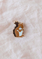 L’épinglette Écureuil MTL de Mimi & August, en brun et blanc, repose sur un tissu beige pâle un accessoire trop mignon, inspiré de Montréal, parfait pour les amoureux des animaux.