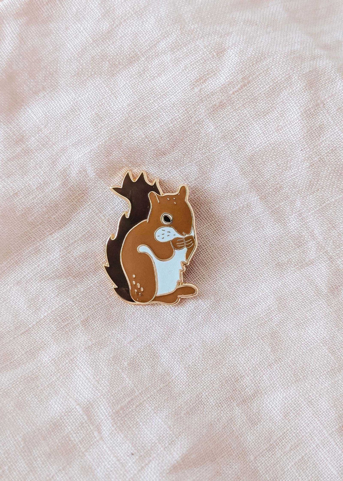L’épinglette Écureuil MTL de Mimi & August, en brun et blanc, repose sur un tissu beige pâle un accessoire trop mignon, inspiré de Montréal, parfait pour les amoureux des animaux.
