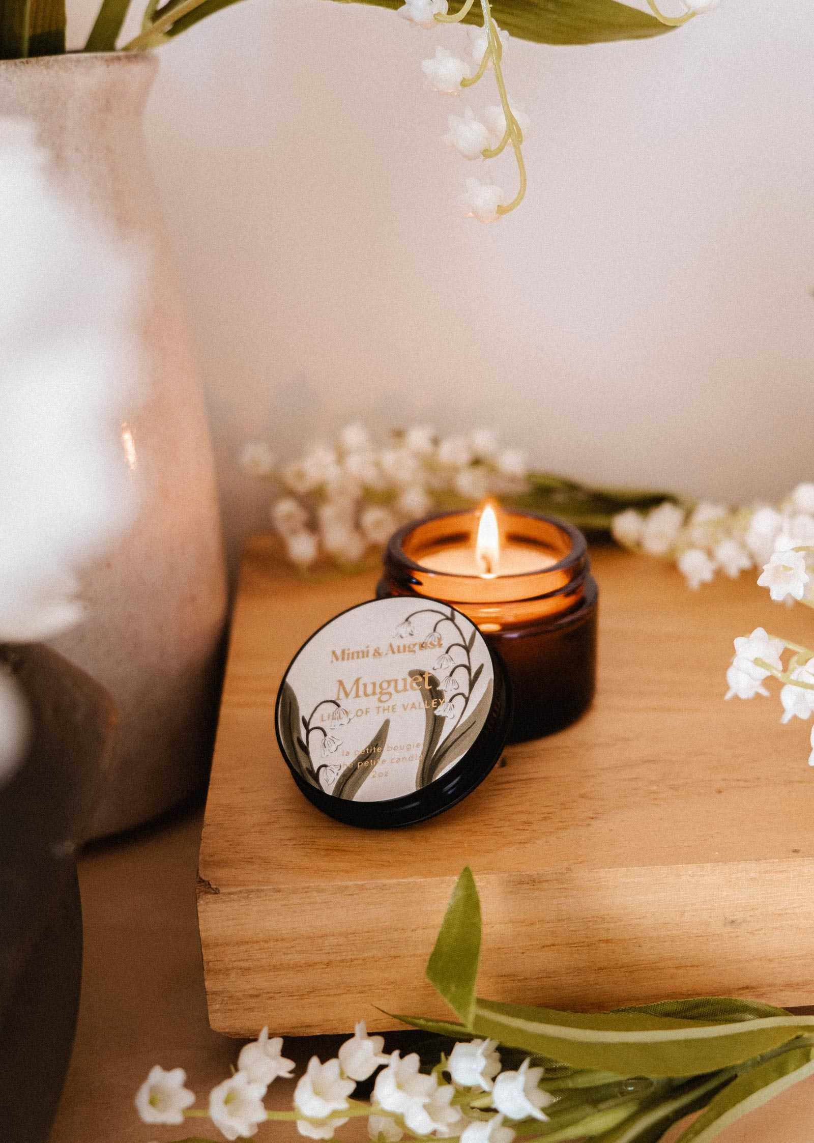 Une bougie de soya Muguet de 2 oz de Mimi & August allumée dans un pot en verre brun repose sur du bois à côté d'un contenant ouvert étiqueté « Muguet », entourée de fleurs blanches et d'un vase en céramique.