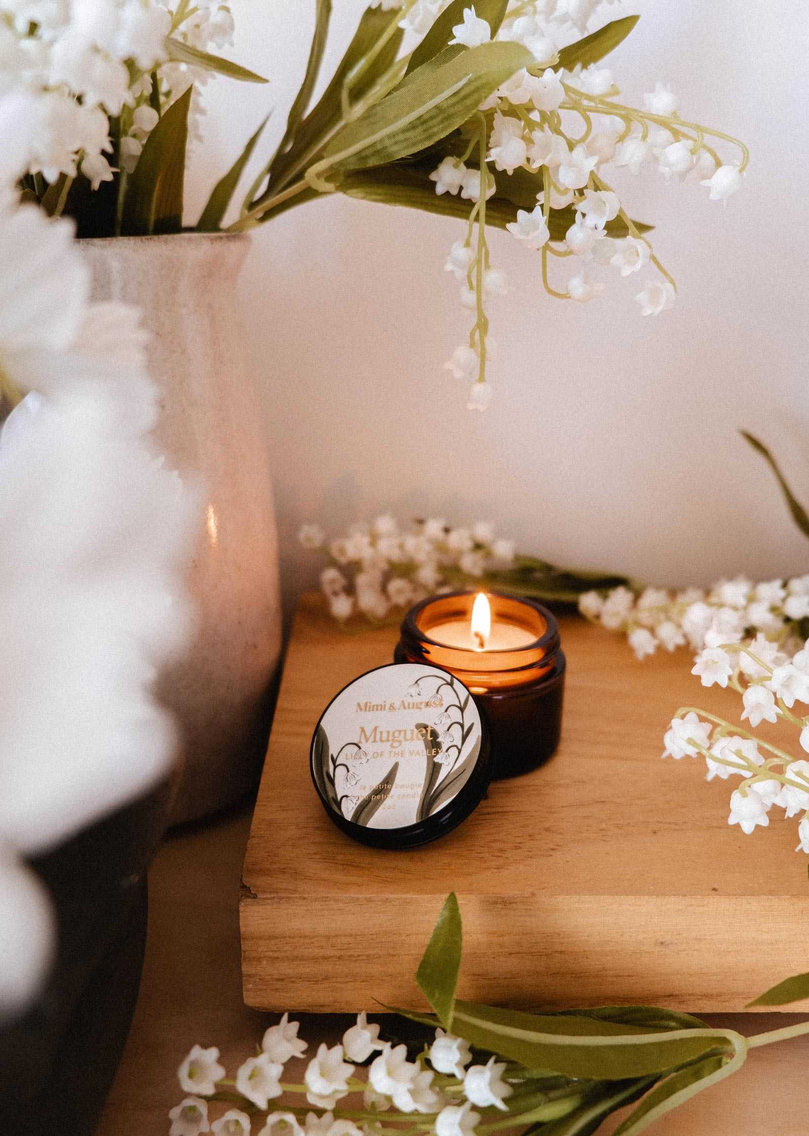 Une bougie de soya Muguet de 2 oz de Mimi & August repose sur du bois parmi des fleurs de muguet et un vase métallique, créant une scène sereine qui met en valeur cette bougie au parfum floral.