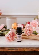 Une petite bouteille d'huile parfumée Musea de Mimi & August repose sur une surface en marbre, avec des œillets roses, une bougie blanche et un livre à proximité.
