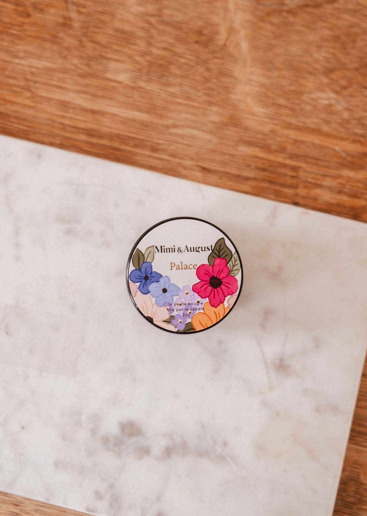 A round tin labeled "Mimi & August" with a colorful floral design sits on a marble surface, showcasing the Palace – 2 oz luxury soy candle, hand-poured in Montreal.