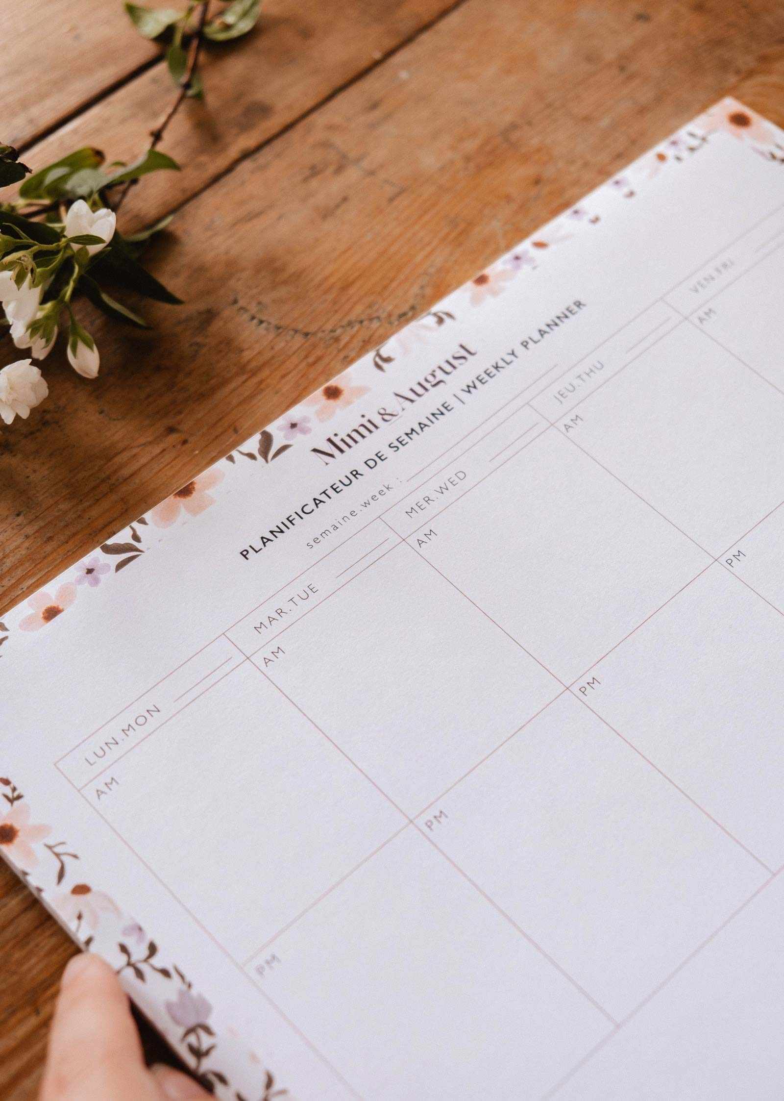 A hand holds the "Petals - Weekly Planner" by Mimi & August, a 50-page French floral planner with blank weekly sections and AM/PM columns, resting beside white flowers on a wooden surface, perfect for organizing your week.