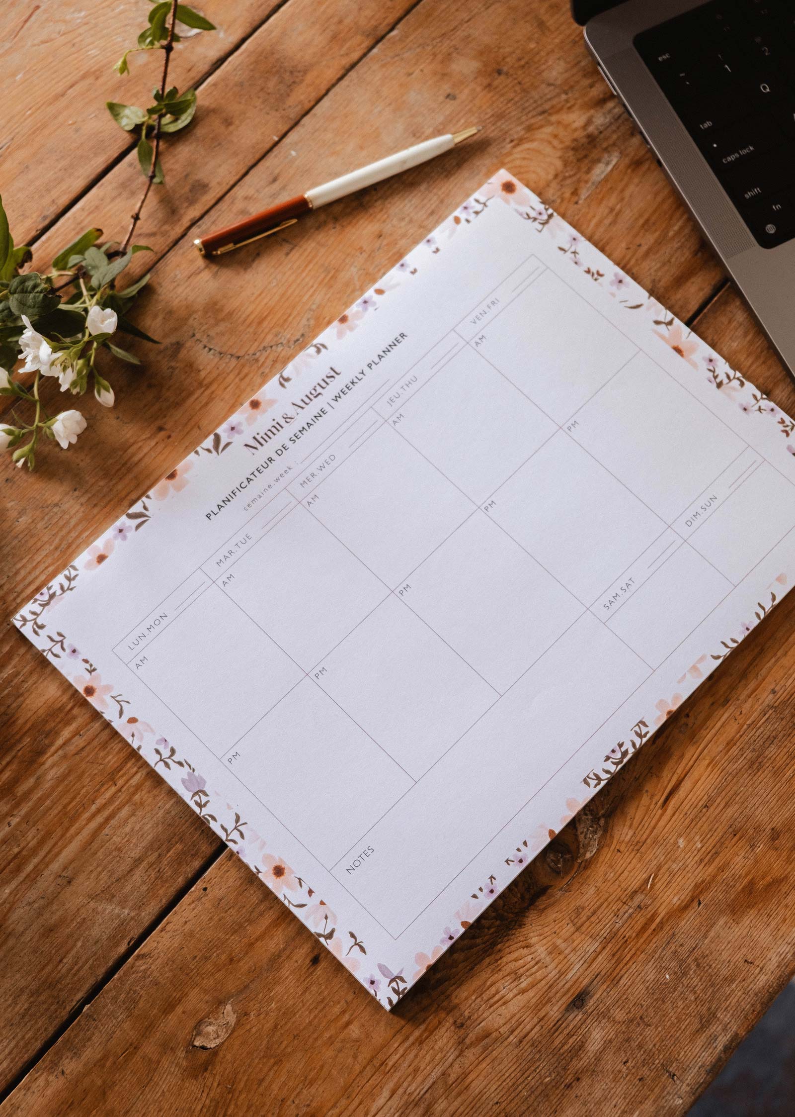 A blank page from the Petals - Weekly Planner by Mimi & August rests on a wooden table beside a pen, a delicate branch with white flowers, and part of a laptop.