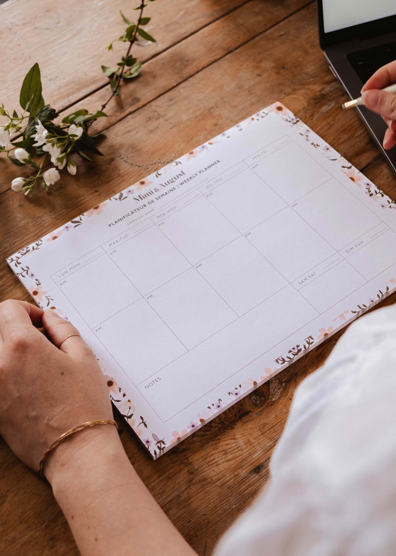 Someone writes on the Mimi & August Petals - Weekly Planner, made from recycled paper, which sits on a wooden table with a sprig of flowers and a laptop close by.