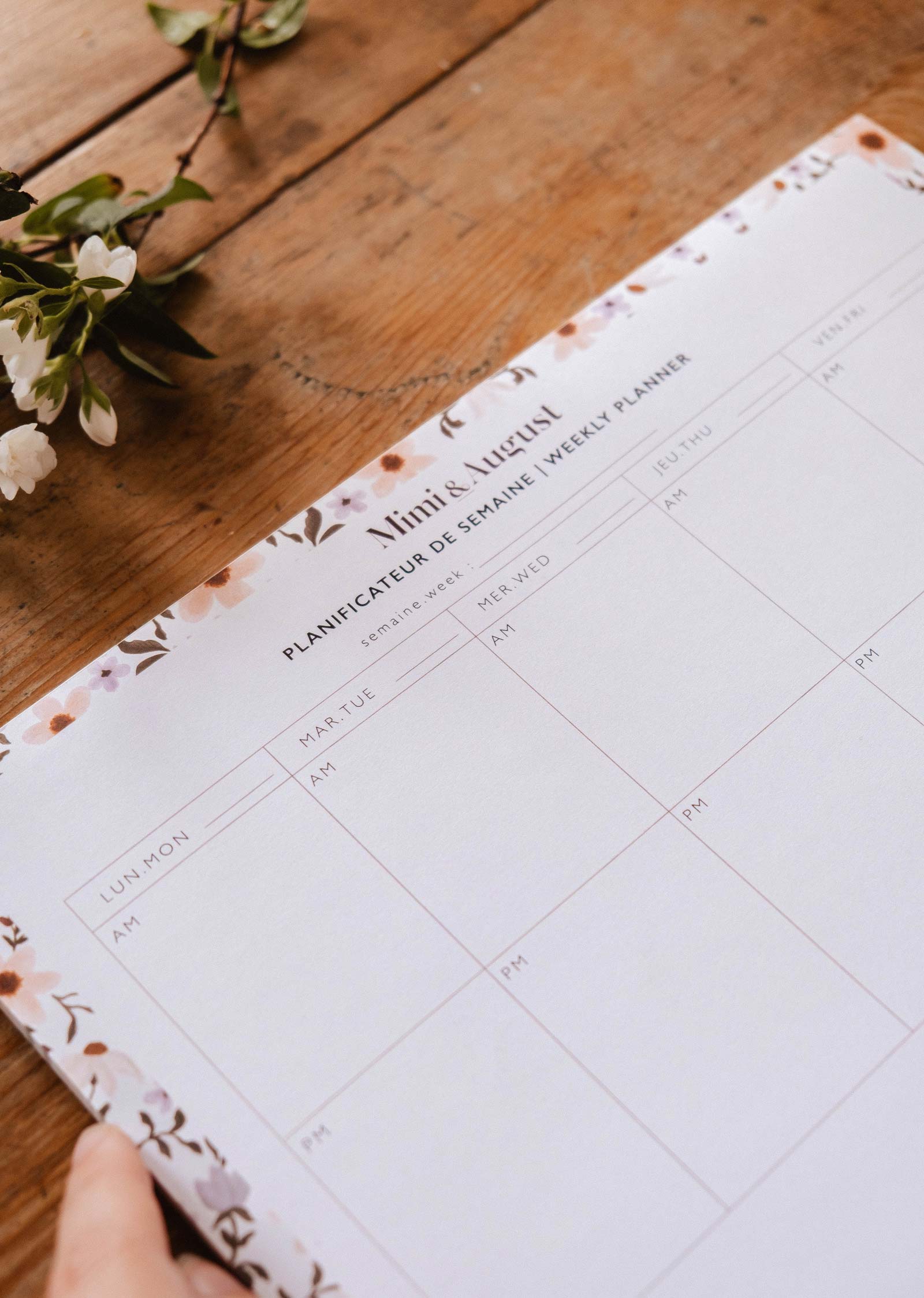 A hand holds the "Petals - Weekly Planner" by Mimi & August, a 50-page French floral planner with blank weekly sections and AM/PM columns, resting beside white flowers on a wooden surface, perfect for organizing your week.