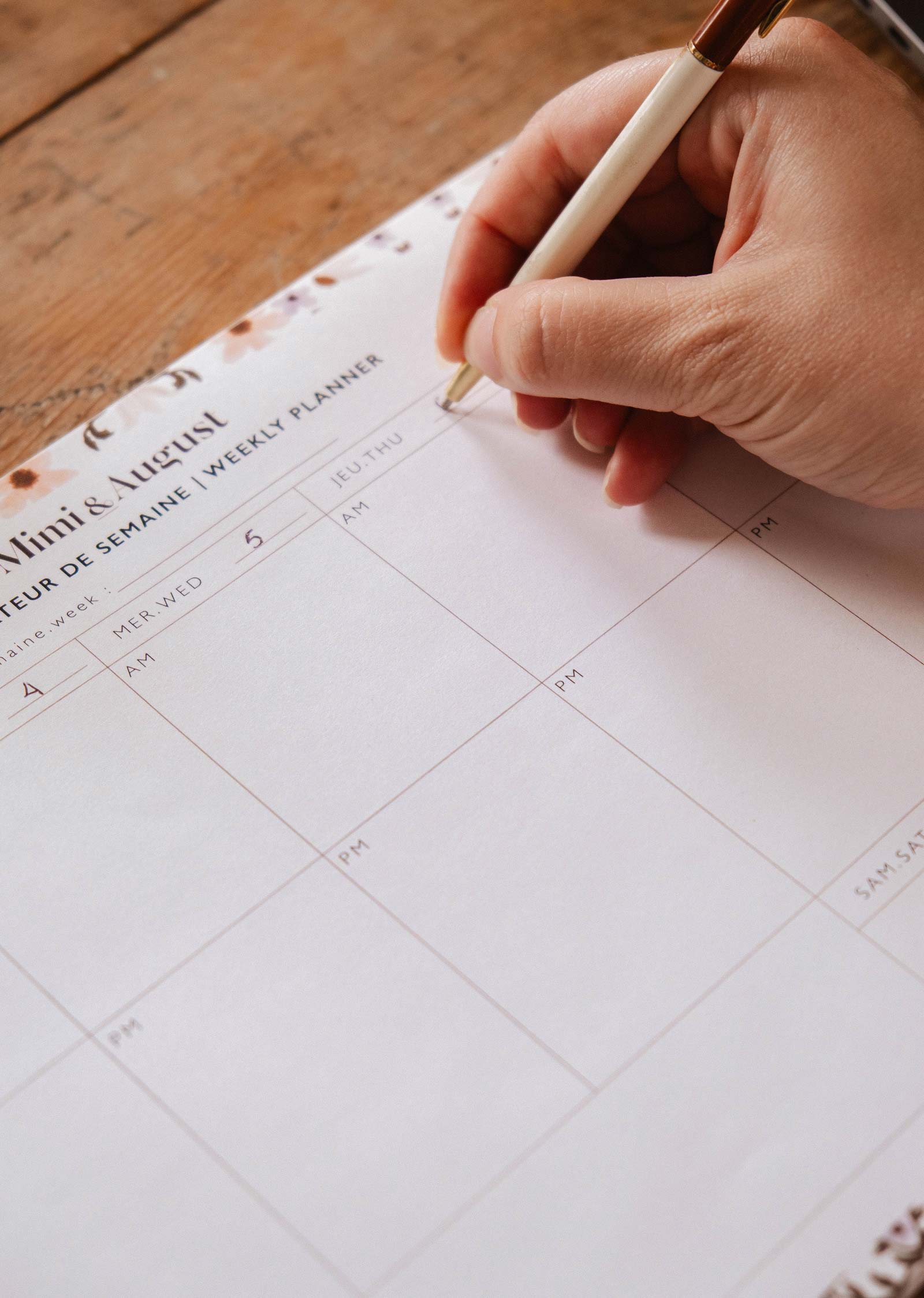 A hand holding a pen writes on a blank Petals - Weekly Planner by Mimi & August, made of recycled paper and set on a wooden surface.