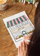 Someone colors a Mimi & August Petit Café Coloring Sheet, featuring a café with striped awnings labeled "Petit Café," surrounded by markers, a candle, and a woven placemat on the table.