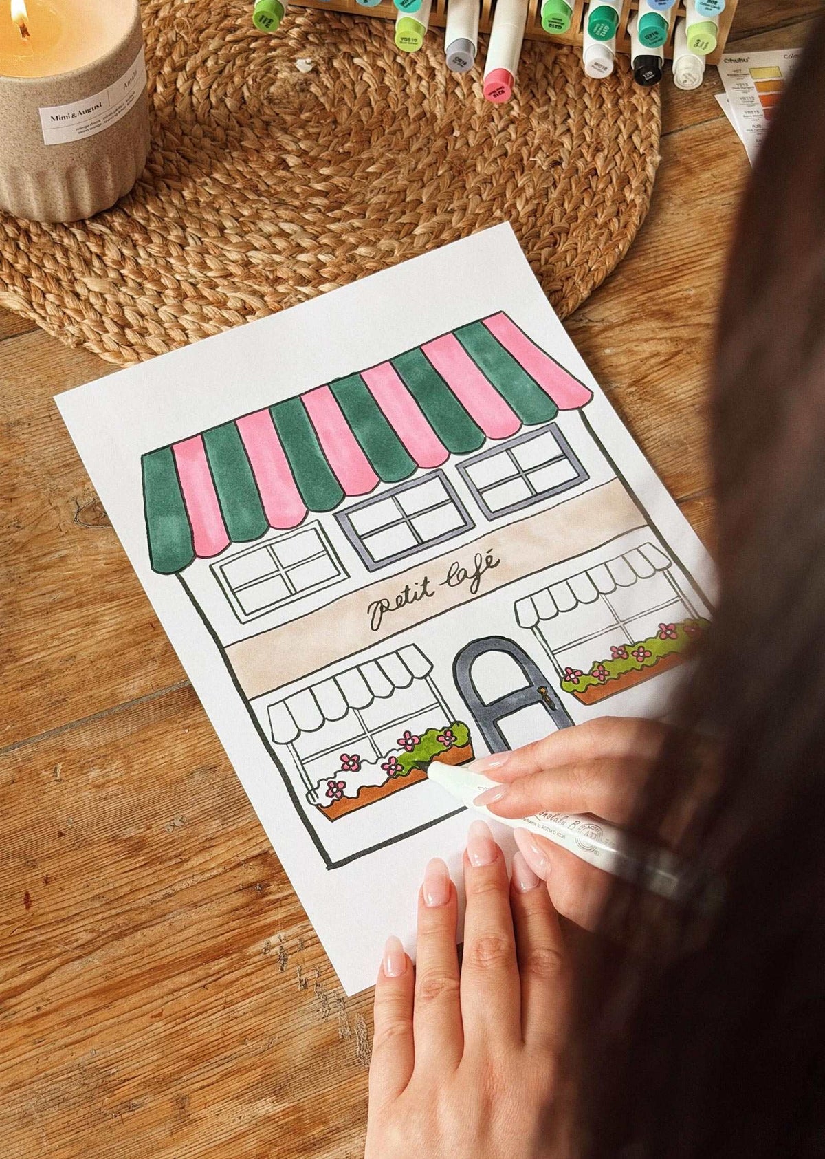 Someone colors a Mimi & August Petit Café Coloring Sheet, featuring a café with striped awnings labeled "Petit Café," surrounded by markers, a candle, and a woven placemat on the table.