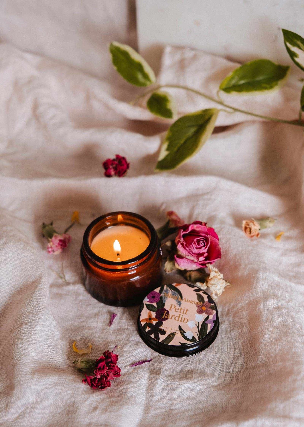 La bougie de soya Petit Jardin 2 oz de Mimi & August, au parfum floral, est coulée à la main à Montréal et repose sur un tissu clair avec un couvercle décoratif en étain et des fleurs séchées et feuilles éparpillées.