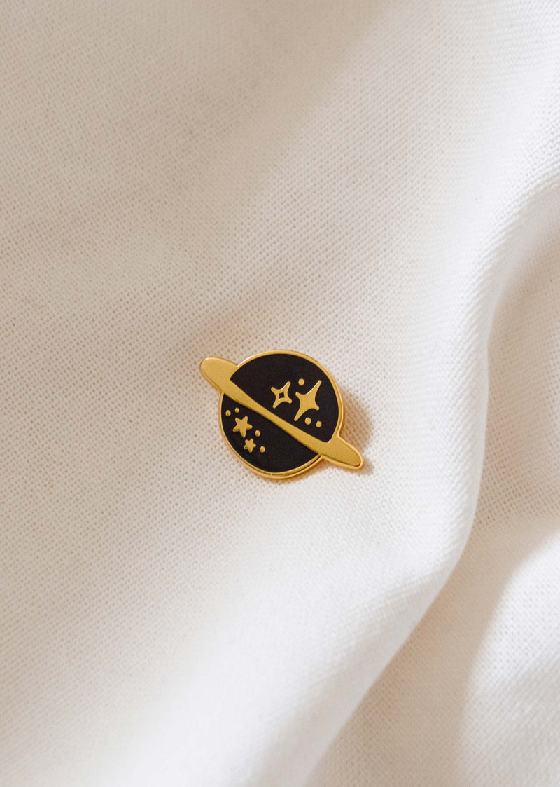 The planet enamel pin by mimi and august on a white shirt.