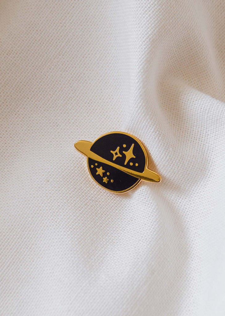 A cosmic black and gold planet stars enamel pin on a white shirt.