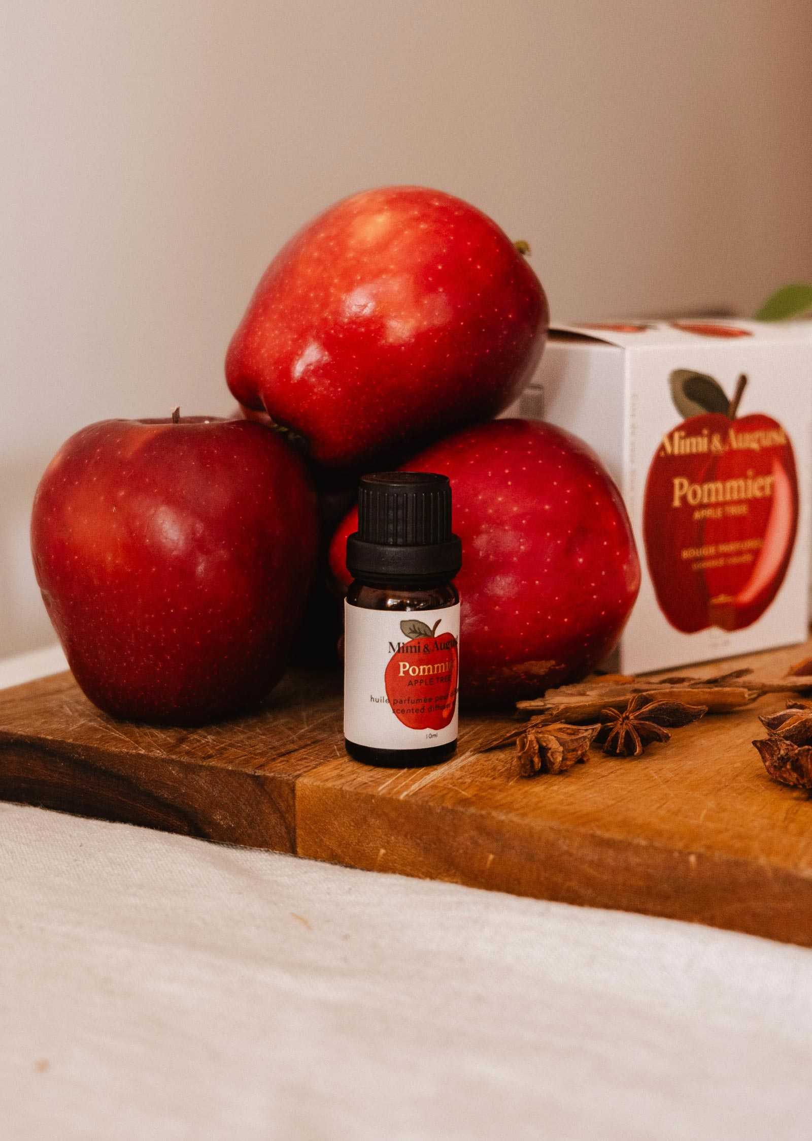 A small bottle of apple cinnamon scented oil, perfect for autumn diffusers, is set on a wooden board in front of three red apples and a box featuring an apple image.