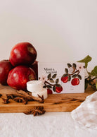 apple cinnamon wax melts cubes - Pommier by Mimi & August rests on a wooden board alongside red apples, two white round wax melts, star anise, and a checkered cloth—perfectly capturing the cozy essence of an autumn home fragrance.