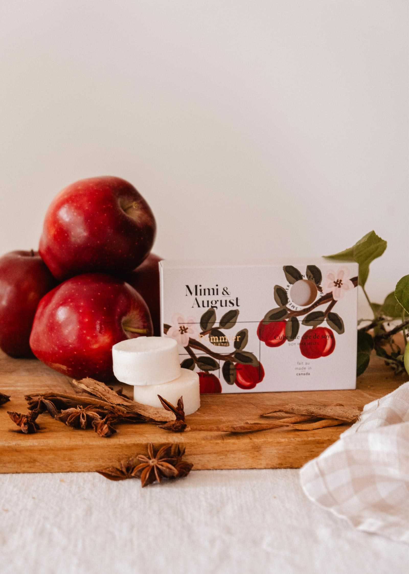 apple cinnamon wax melts cubes - Pommier by Mimi & August rests on a wooden board alongside red apples, two white round wax melts, star anise, and a checkered cloth—perfectly capturing the cozy essence of an autumn home fragrance.